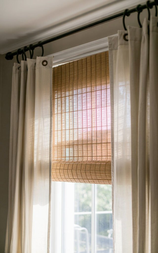 Bamboo roman shade layered under white linen curtains creating elegant window treatment for bedroom